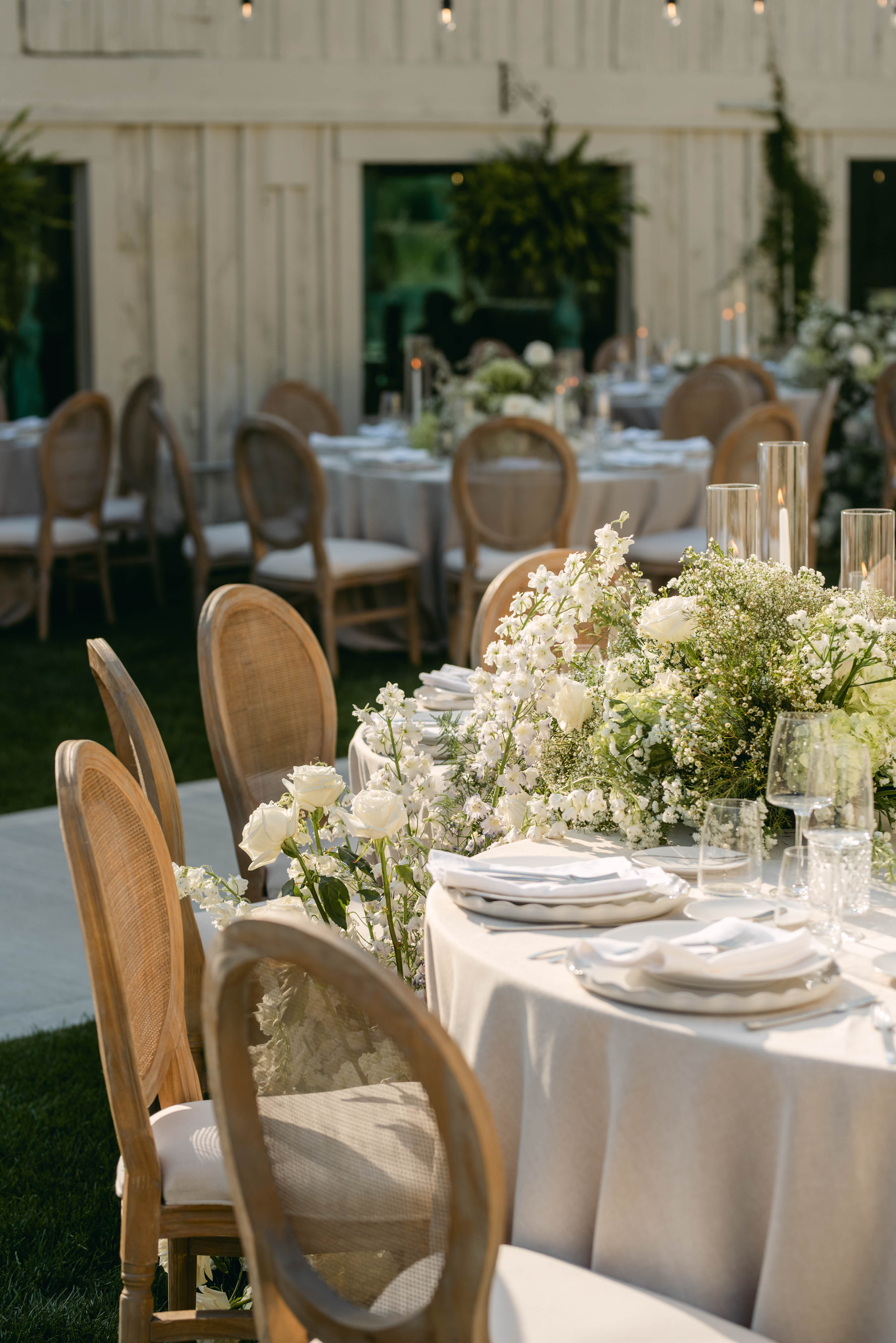 Wedding tablescape with cascading white florals, taper candles, and cane chairs against the barn